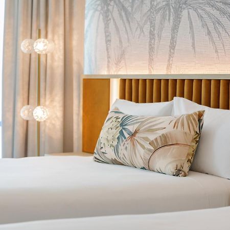 Amora Hotel Бризбейн Екстериор снимка The photo shows a stylish hotel room featuring two neatly made beds with white linens. Each bed has several pillows, one of which has a decorative cover with a tropical plant pattern. Behind the beds is a modern headboard, and the wall is decorated w