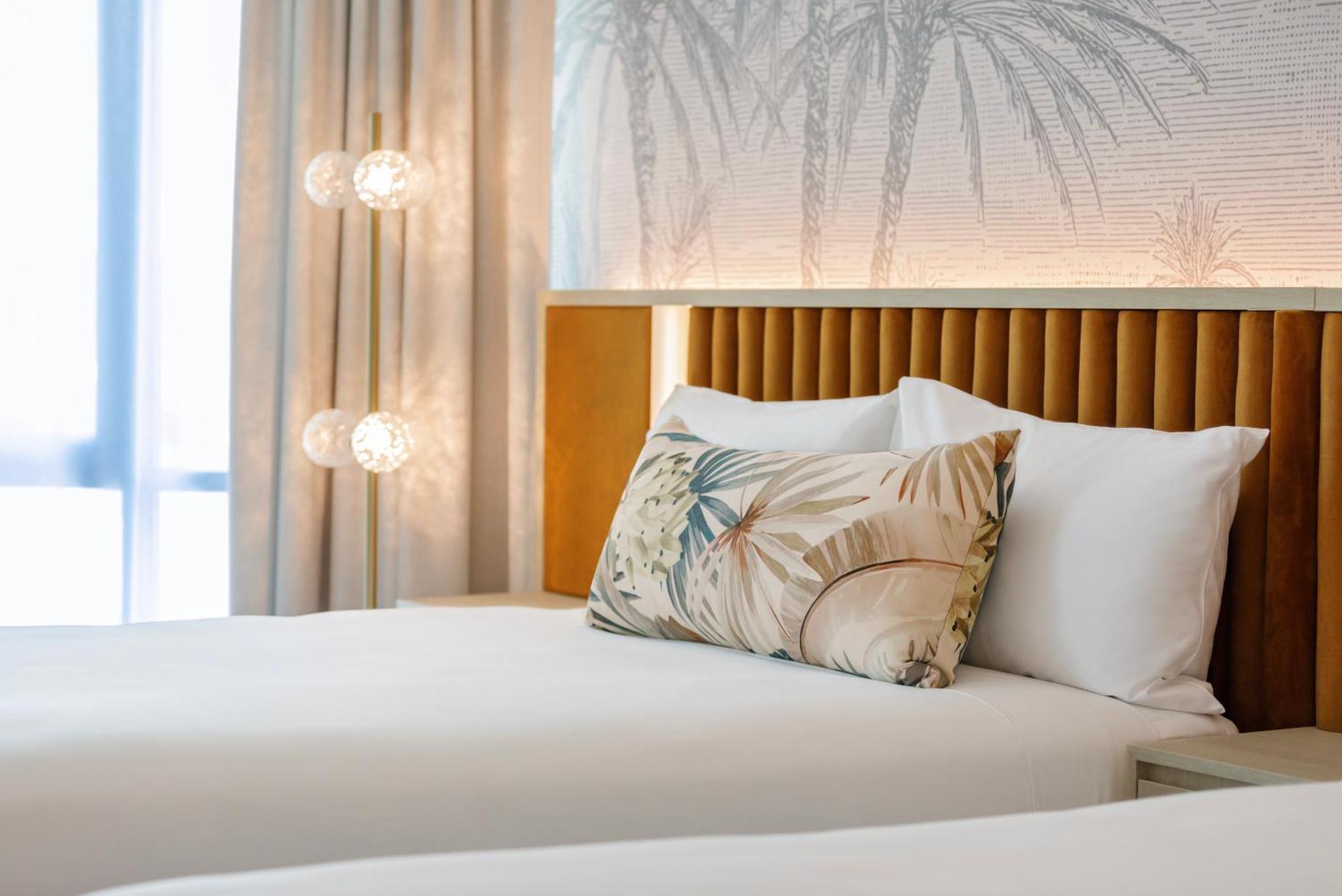 Amora Hotel Бризбейн Екстериор снимка The photo shows a stylish hotel room featuring two neatly made beds with white linens. Each bed has several pillows, one of which has a decorative cover with a tropical plant pattern. Behind the beds is a modern headboard, and the wall is decorated w